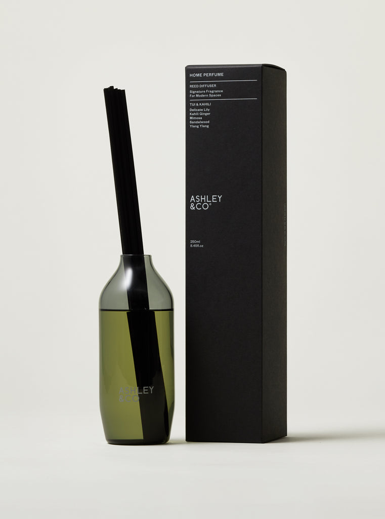 ASHLEY & CO home fragrance reed diffuser bottle with a black reed and olive-green liquid, standing next to its tall black packaging box on a light gray background.