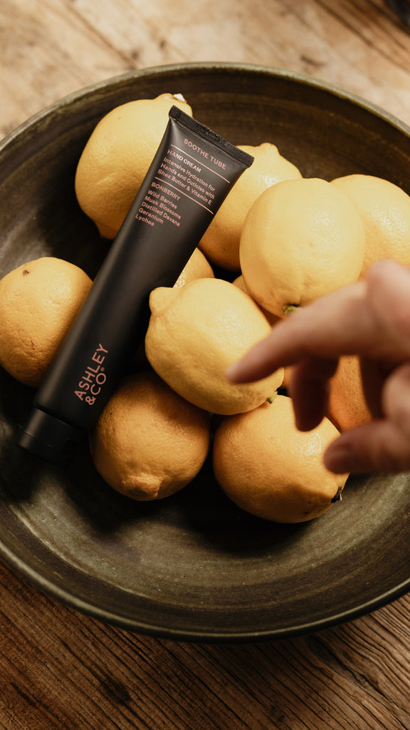 A black tube of Ashley & Co Vitamin C Serum for Sensitive Skin resting on a dark ceramic bowl filled with lemons, with a person's hand pointing toward the product.