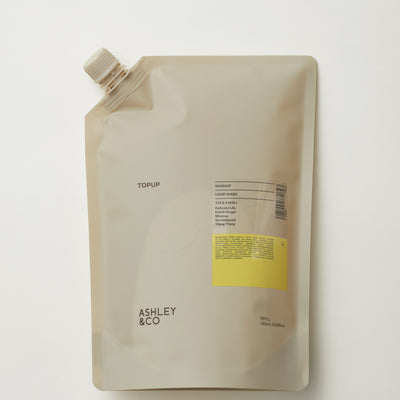 A stand-up resealable pouch package with a small spout cap, shown on a clean white background. The pouch is a warm light beige/taupe color with black text reading 'TOPUP' and 'ASHLEY & CO', plus a small yellow label block.