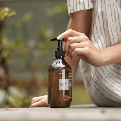 A person in a white and gray striped shirt is shown from the side, pressing the black pump dispenser of an amber glass bottle placed on a wooden table outdoors.
