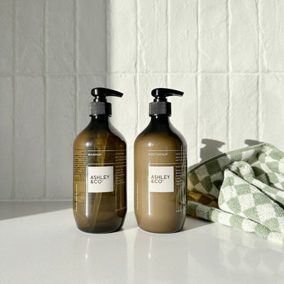 Two amber glass pump bottles with black dispensers and white labels reading ASHLEY & CO, standing on a white surface against a white tiled wall, with a green-and-white checkered towel partly visible on the right.