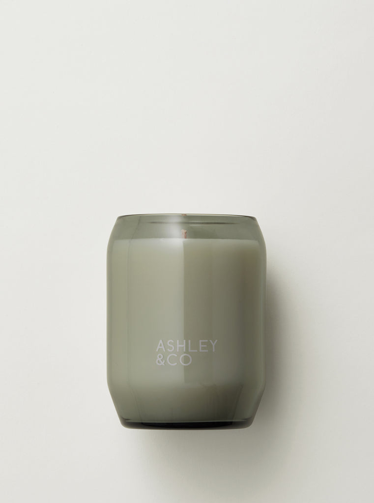 A smoky gray glass candle jar with a matte finish and the brand name 'ASHLEY & CO' embossed on the front, set against a clean off-white background.